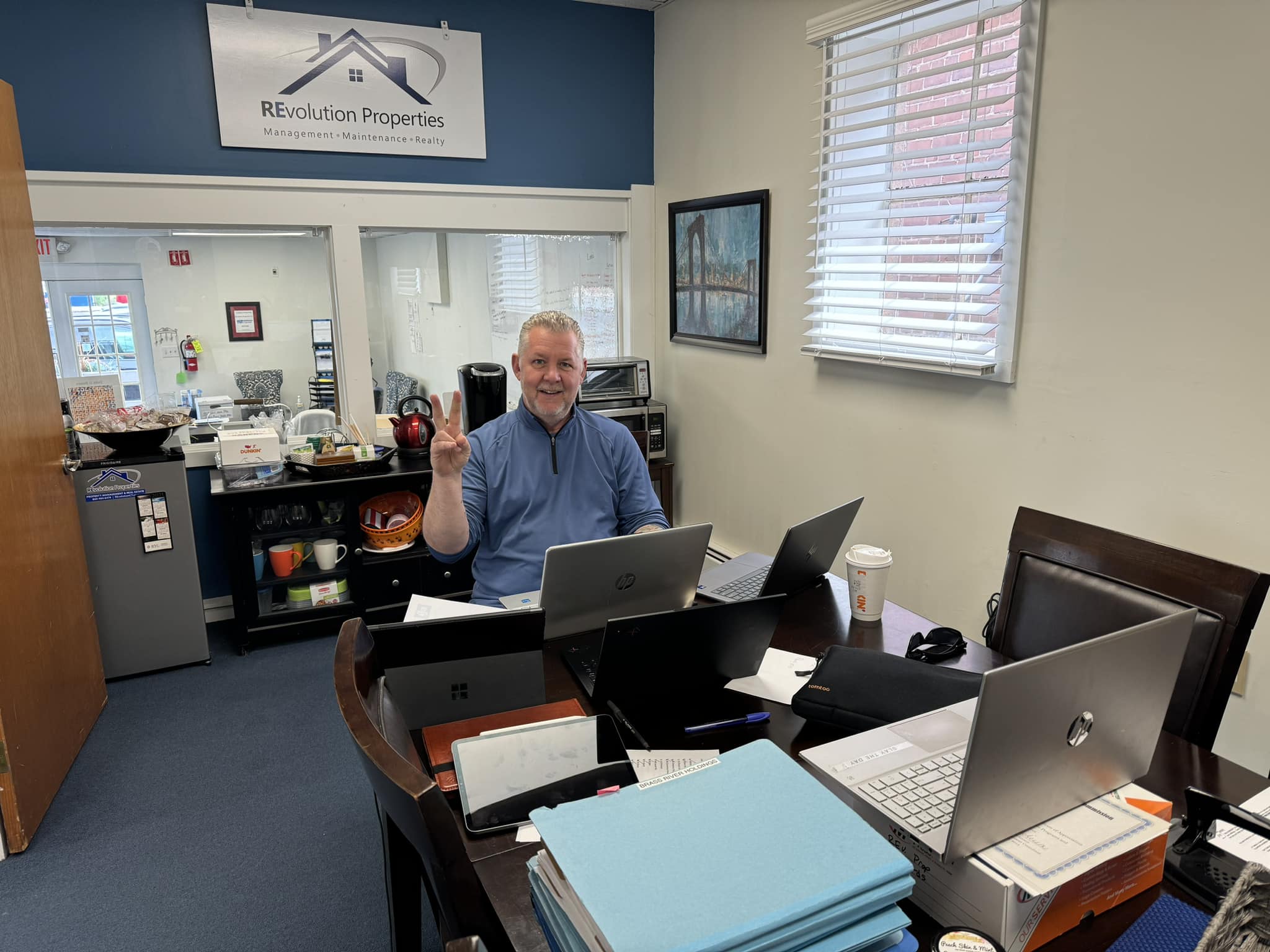 Smiling man sitting at a desk in a commercial property management office, holding up a peace sign and a coffee mug. Multiple laptops, documents, and office supplies are spread across the table. A company sign reading 'REvolution Properties' is visible on the wall behind him.